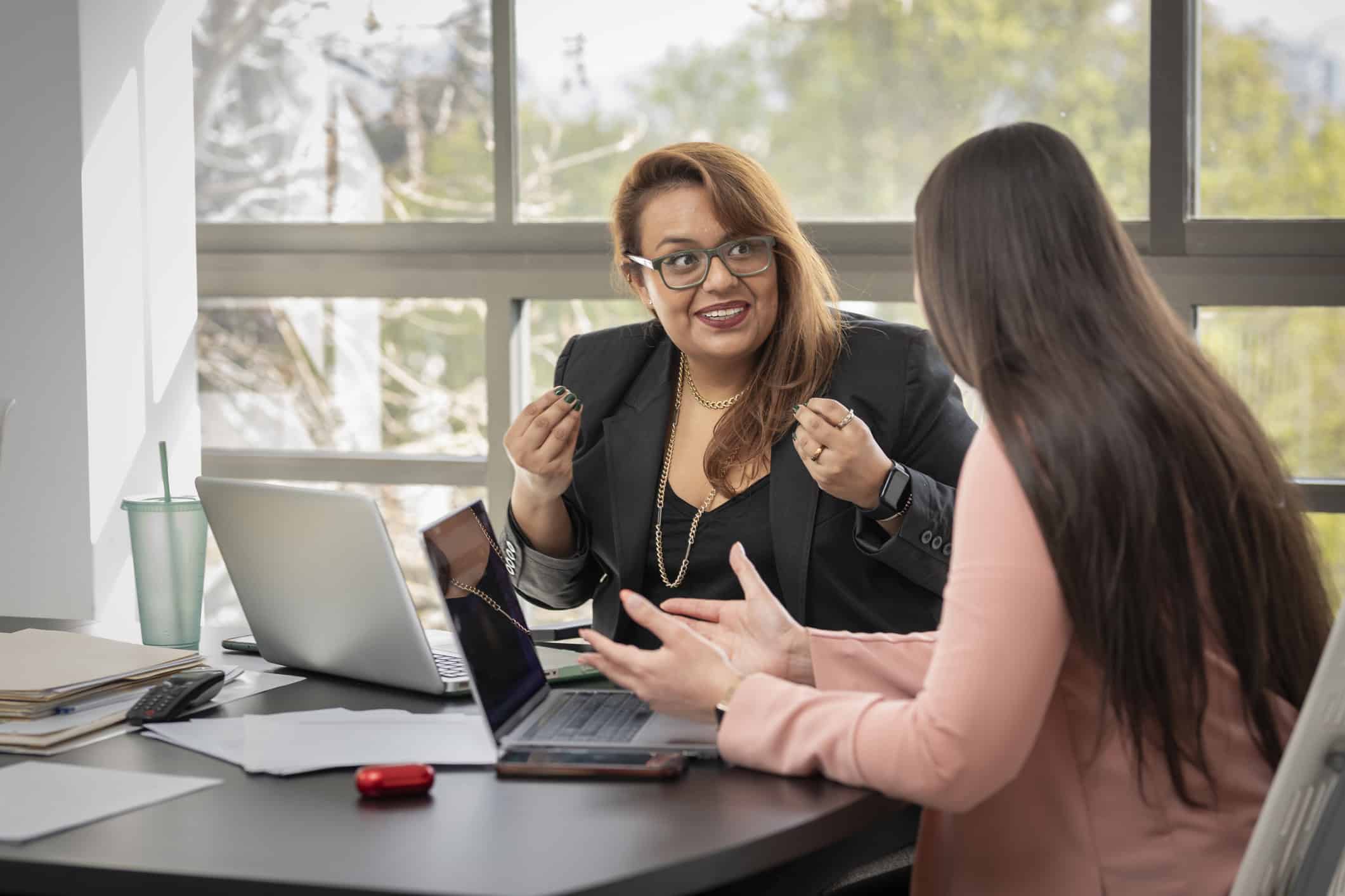 Two women at the office talking to one another. The women are from 2 different backgrounds, they're both smiling.