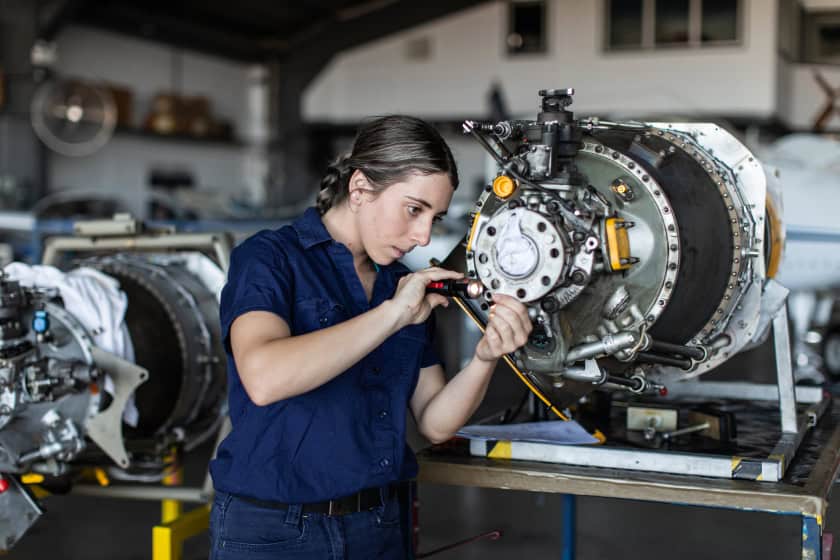 Photo of a young engineer working. The engineer is a young blonde woman, her expression is focused. She is working on a large steel engine of some sort.